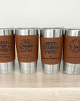 Four stainless steel tumblers with brown leather wraps and engraved text on a light wooden surface.