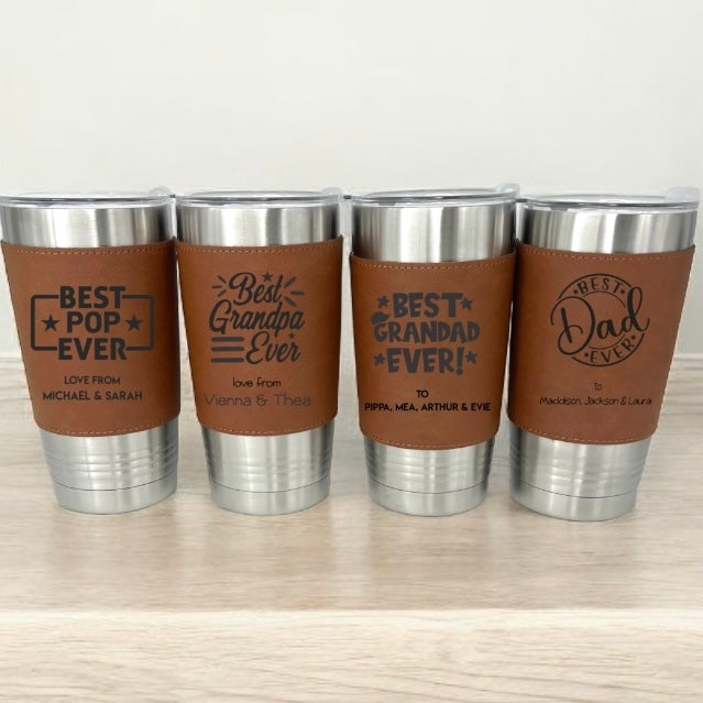 Four stainless steel tumblers with brown leather wraps and engraved text on a light wooden surface.